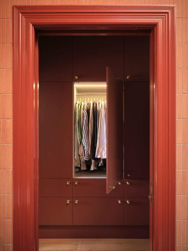 A built-in wardrobe and dressing storage unit viewed through an ornate red-painted doorway. The cabinetry is finished in deep reddish-brown, with brass knobs and a central open hanging section illuminated by warm integrated lighting. The surrounding wall and heavily moulded door trim are terracotta-red, while a timber floor is visible at the base.
