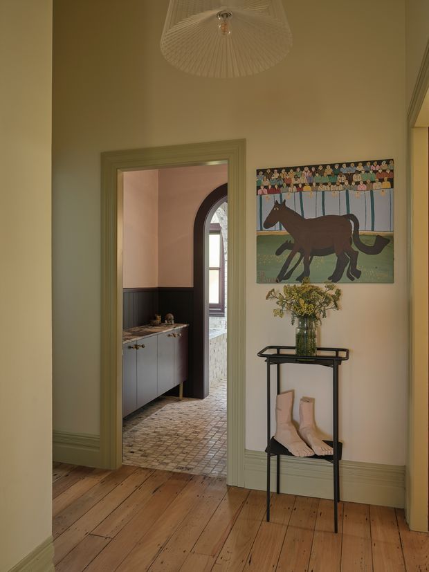 A warmly lit traditional hallway or entryway with wide timber floorboards, pale green painted walls and matching trim, and a decorative pendant light. A narrow black console displays a vase of flowers and boots beneath a colourful horse artwork. Through the open doorway is a small utility or pantry-like space with dark lower cabinetry, patterned tiled flooring, and an arched opening.