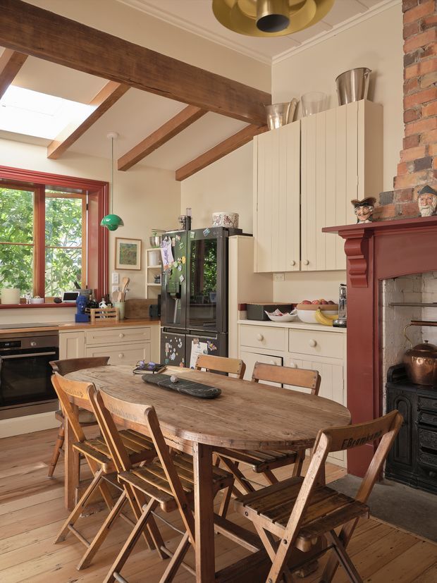 A warm, traditional kitchen-dining space with cream cabinetry, a large rustic timber dining table, folding timber chairs, exposed timber ceiling beams, and a red-painted brick fireplace with a dark cast-iron stove. The room includes a black refrigerator, timber flooring, a red-trimmed window, and a brass pendant light. The overall character is farmhouse-inspired and lived-in.