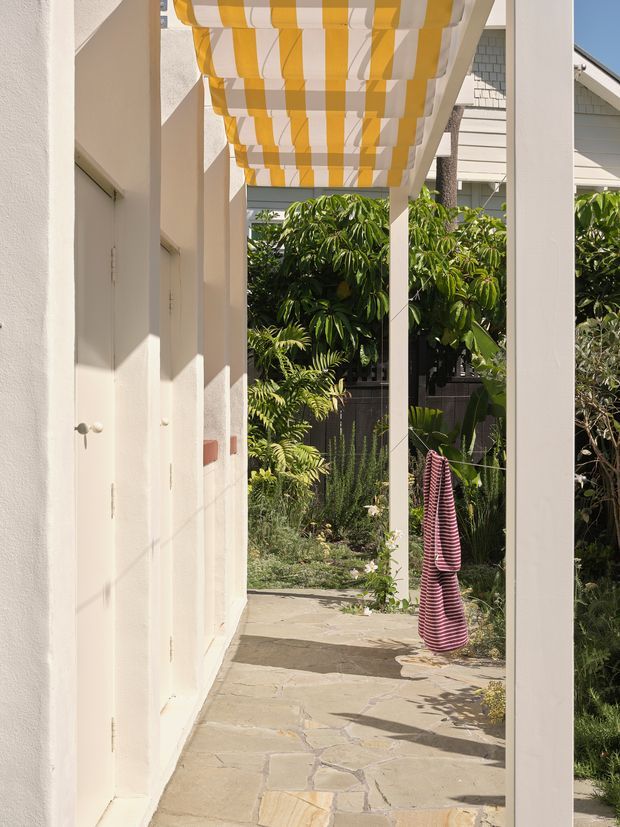 A bright covered outdoor passage beside a white rendered building, supported by simple white posts and topped with a striped yellow-and-white fabric awning. The walkway has irregular natural stone paving and opens onto a lush planted garden with tropical foliage. A striped textile hangs from a clothesline, adding a casual residential outdoor character.