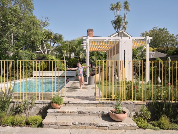 Sunny landscaped residential backyard with a swimming pool enclosed by bright yellow metal fencing. Stone steps lead to a paved pool terrace and a white pergola with a yellow-and-white striped retractable canopy. Tropical and subtropical planting surrounds the space, with a child near the pool entrance.