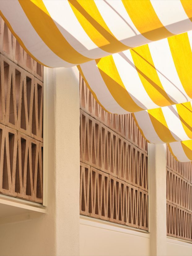 A sunlit architectural courtyard or passage with cream stucco walls, vertical structural columns, and decorative terracotta breeze-block screens. Yellow-and-white striped fabric canopies create shade overhead, producing a warm, patterned atmosphere.