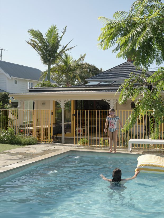 Sunny residential outdoor area with a swimming pool in the foreground, a covered veranda with decorative arches and gold-toned metal fencing, and a light-coloured house with corrugated metal pitched roofs behind it. Tropical planting surrounds the pool and veranda.
