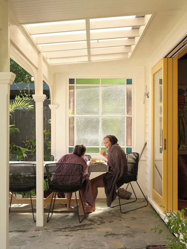 A bright, enclosed verandah or sunroom with a translucent roof, white weatherboard walls, decorative timber posts, and large textured-glass windows. A small dining table and black wire-frame chairs create a casual seating area, with folding timber-framed doors opening into the adjacent interior. Natural greenery is visible outside, and the irregular stone floor reinforces the indoor-outdoor character.