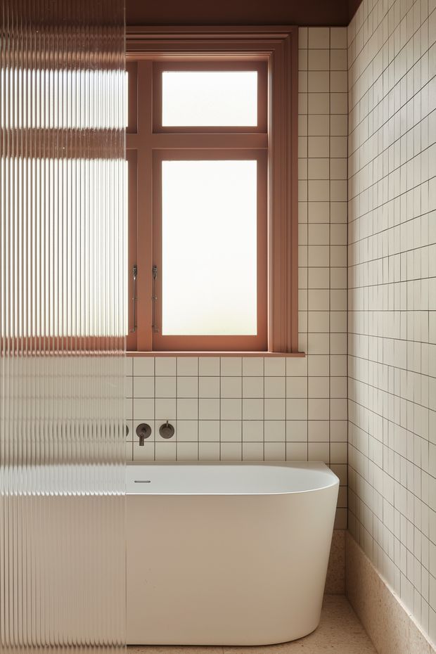 A warm, compact bathroom featuring a freestanding white bathtub, cream square wall tiles with dark grout, a terrazzo-like floor, and a timber-framed frosted window. A ribbed translucent glass partition partially screens the tub on the left. The room has a refined, contemporary appearance with terracotta-brown painted window and ceiling trim.