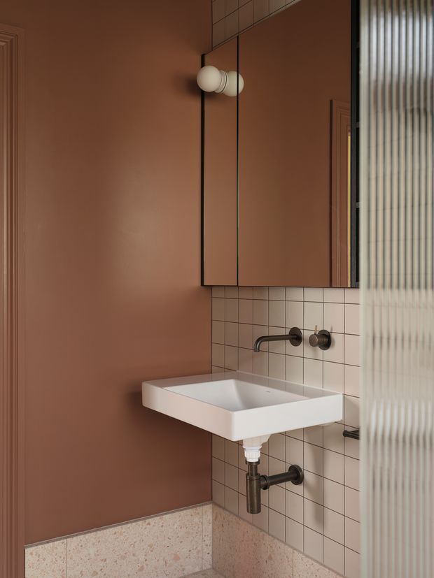 Compact contemporary bathroom vanity with a wall-mounted white rectangular basin, exposed dark metal plumbing, wall-mounted faucet, dusty terracotta painted walls, pale square tiled splashback, and terrazzo-style lower wall tiles. A mirrored cabinet with globe lighting is mounted above the basin, while ribbed glass partially screens the right side.