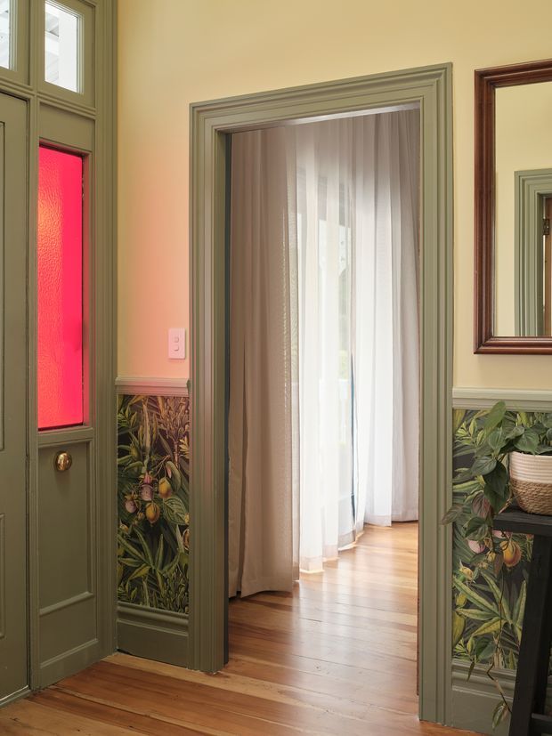 A richly detailed traditional entryway with olive-green painted timber paneling and trim, a glazed front door featuring a vivid red textured glass panel, and decorative botanical wallpaper wainscoting. An open doorway leads into a brighter room with sheer curtains and timber flooring. A framed mirror and potted plant are visible on the right.