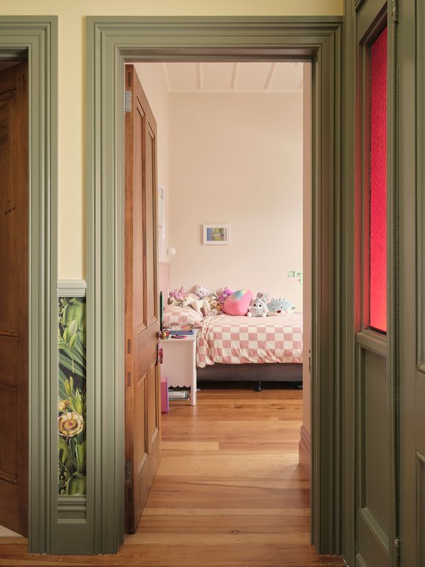View through a richly trimmed interior doorway into a child's bedroom. The bedroom has warm timber flooring, pale pink walls, a pink-and-white checked bedspread, plush toys, and a timber bed. The foreground hallway features muted sage-green painted moulded door and wall trim, decorative floral tile or painted panels, and a red translucent glass sidelight. The scene has a heritage character with layered architectural detailing and a warm, playful bedroom interior.