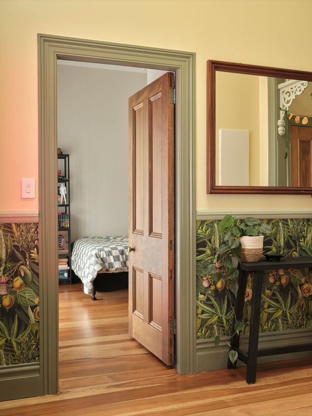 A richly detailed traditional hallway with a timber floor, ornate botanical wall covering below painted dado trim, and muted green decorative door and architrave mouldings. A timber-panelled door stands open to a bedroom with a patterned bedspread and timber floor. A large framed mirror, console table, and potted plant decorate the hallway.