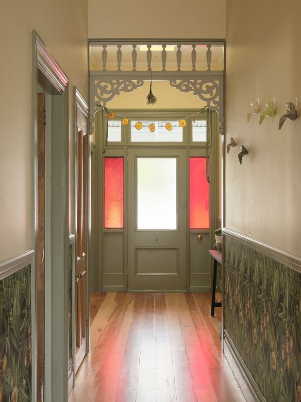 A narrow, richly detailed period-style hallway leading to a glazed front door. The space features warm timber floorboards, muted green painted joinery and door, patterned botanical wall panelling, decorative carved fretwork, a balustraded transom opening, stained-glass side panels, and wall-mounted decorative objects. Strong red and amber light filters through the coloured glass, creating a warm historic atmosphere.