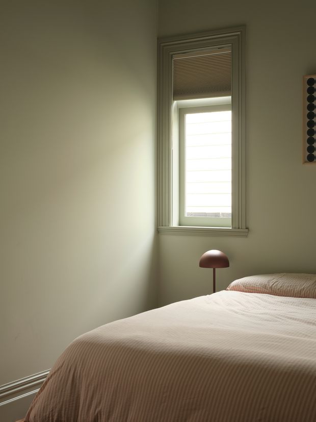 A quiet, softly lit bedroom with a neatly made bed covered in a warm beige textured bedspread. A small rounded dome table lamp sits beside the bed, while a tall window with layered trim and a partially lowered blind provides diffused daylight. The muted neutral palette and minimal furnishings create a calm, understated atmosphere.