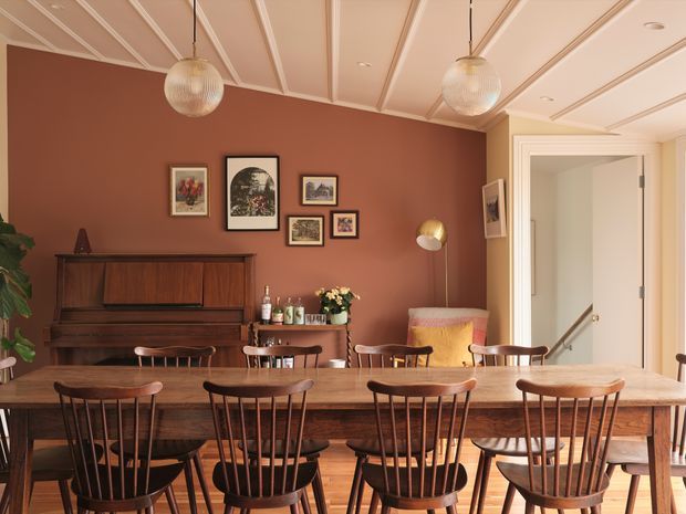 A warm, characterful dining area with a long solid-wood dining table and multiple spindle-back wooden chairs in the foreground. The room features a dark timber upright piano, a small timber sideboard, framed artwork on a terracotta-brown wall, a blush upholstered armchair, globe pendant lights, and a white coffered or panelled ceiling. An open doorway and stair handrail are visible at the right.