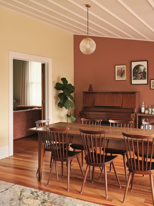 Warmly styled dining room with a long solid-wood dining table and matching spindle-back wooden chairs, an upright piano against a terracotta feature wall, framed artwork, a bar cabinet, a pendant light, and decorative painted ceiling mouldings. Timber flooring extends toward a connected living room visible through the doorway.