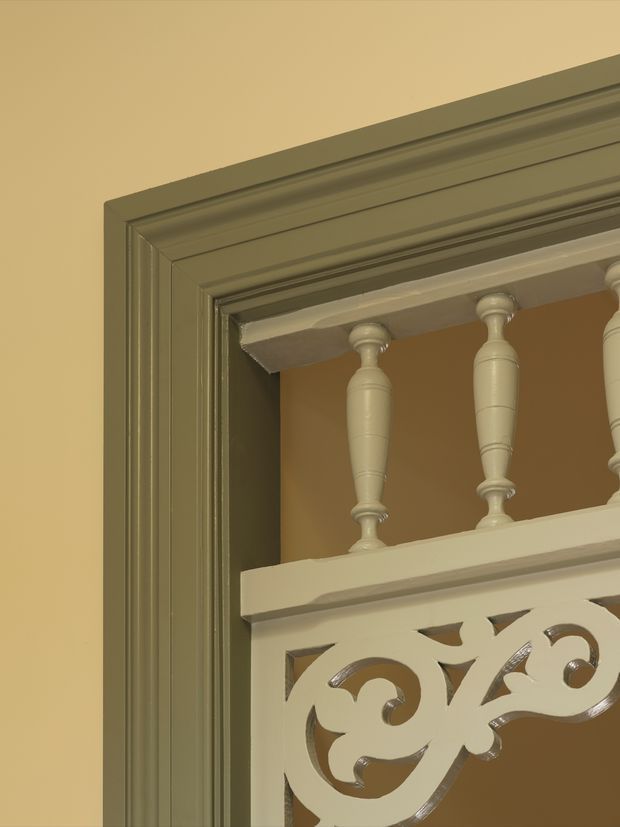 Close-up architectural detail of an ornate traditional interior opening, featuring layered dark olive-green moulded trim, cream-painted turned balusters, and decorative scrollwork balustrade panels against a warm beige wall.