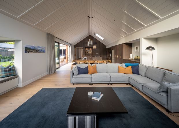 A spacious open-plan living room with a large L-shaped light grey sectional sofa, dark coffee table, and deep blue area rug. The room opens into a dining and kitchen area, with warm timber flooring, dark timber kitchen cabinetry, a pitched white-lined ceiling with exposed beams, roof skylight, pendant lights, and extensive glazing leading outdoors. Neutral walls and timber finishes create a contemporary, warm interior.