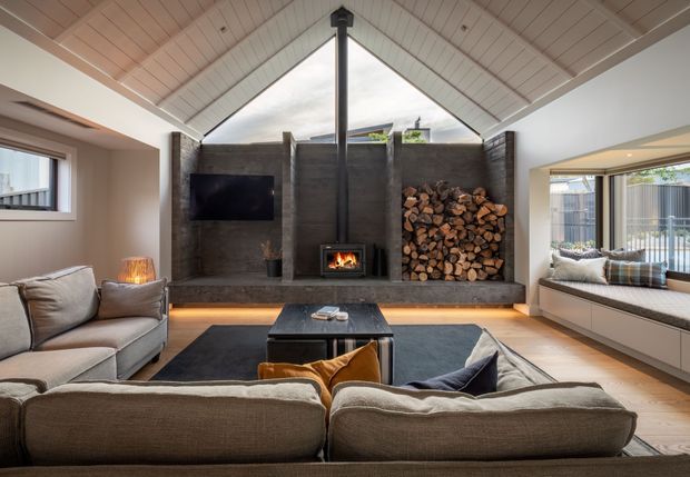 A warm contemporary living room centred on a dramatic double-height gabled ceiling and a dark concrete fireplace wall. The wall incorporates a built-in television, suspended black flue, wood-burning fireplace and stacked firewood niche, with concealed warm underlighting. Multiple sofas and armchairs surround a low dark coffee table, while built-in window seating and large glazed openings bring in daylight. The space combines pale timber ceiling boards, neutral upholstery and dark masonry-like finishes for a refined, cosy atmosphere.