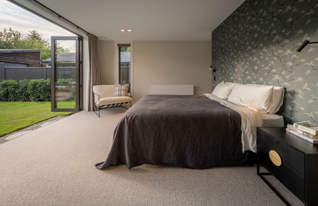A contemporary bedroom with a large upholstered bed dressed in dark charcoal bedding, positioned against a botanical feature wallpaper wall. The room has soft neutral walls, plush light carpeting, integrated bedside joinery, a small upholstered lounge chair, and full-height glazing with a glass door opening to a landscaped garden. Recessed ceiling lights and restrained furnishings create a calm, refined atmosphere.