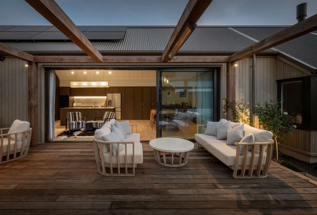 A covered timber deck and outdoor lounge opening directly into a contemporary home. The space features exposed timber beams, dark timber decking, light upholstered lounge seating, a round coffee table, warm architectural lighting, and large glazed sliding doors revealing an interior kitchen and dining area. The surrounding building has dark vertical cladding and corrugated pitched roofing.