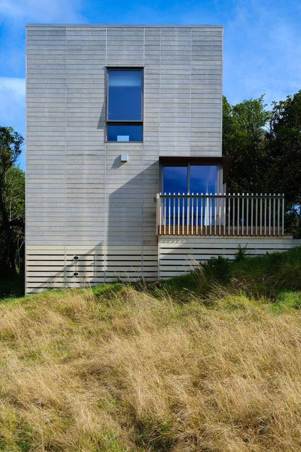 A compact modern multi-storey house viewed from outside, set among long grass and trees. The facade is clad in pale grey horizontal weatherboard-style boards with a simple rectangular form, narrow dark-framed windows, and a projecting balcony or terrace with a vertical metal balustrade. Bright blue sky and surrounding vegetation contrast with the restrained contemporary exterior.