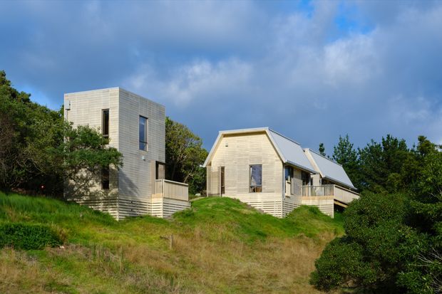 Exterior photograph of two light-coloured timber-clad residential buildings set on a grassy, landscaped sloping site. The left structure is a compact modern two-storey rectangular volume with narrow vertical windows, while the right is a smaller single-storey gabled house with a pitched metal roof, timber deck and simple rectangular glazing. Dense vegetation surrounds the buildings beneath a partly cloudy sky.