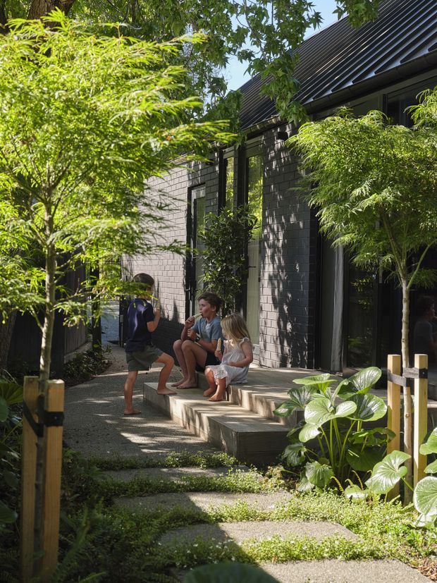 A shaded garden walkway leads to a contemporary dark brick-clad building with a steeply pitched dark metal roof and tall glazed openings. Timber-edged concrete steps form a small entry terrace, surrounded by lush layered planting. Three people sit and stand on the steps, creating a welcoming residential atmosphere.