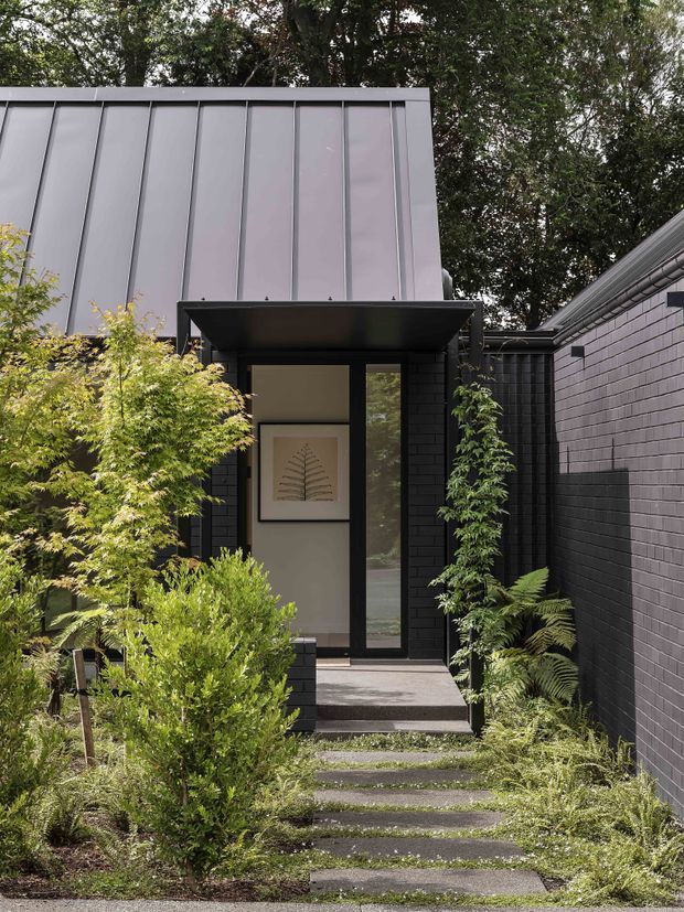 A contemporary residential entrance approached through a lush, landscaped garden. The facade combines dark brick walls with a tall standing-seam metal-clad upper volume and a deep black canopy over a narrow glazed entry door. Stepping stones lead through layered planting, ferns, and shrubs toward the front threshold. The composition has strong vertical lines, restrained dark tones, and a sheltered courtyard-like feel.