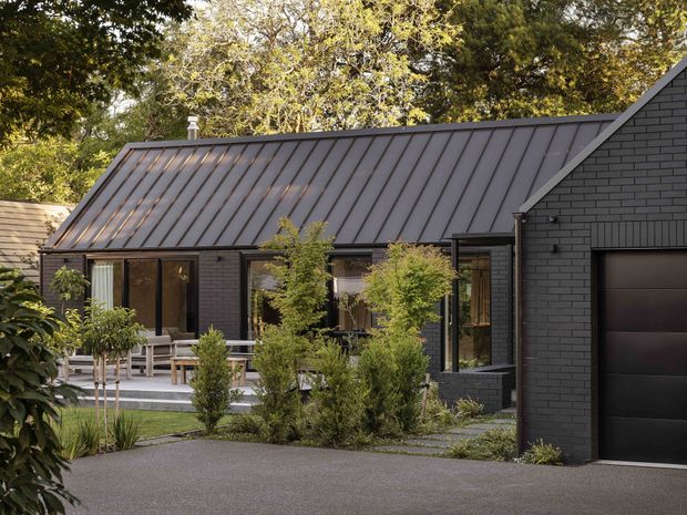 Exterior view of a contemporary dark-clad residential building with a steep standing-seam metal roof, black brick walls, large glazed openings, landscaped planting, paved driveway, and an attached garage. The composition highlights the roofline and facade against mature trees.