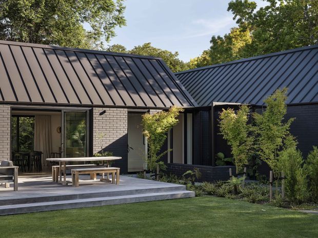 Exterior view of a contemporary single-storey home arranged around a landscaped courtyard or patio. The building has dark standing-seam metal pitched roofs, dark painted brick walls, large glazed sliding doors, and a broad stepped concrete terrace furnished for outdoor dining. Mature trees and lawn frame the composition.