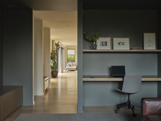 A refined home office nook occupies the right side of the image, with a long built-in desk, open shelving, framed artwork, a laptop and a grey swivel chair. The office opens onto a warm timber-floored hallway leading toward a softly furnished living area. Dark charcoal walls and cabinetry contrast with pale neutral surfaces, while a leather armchair is partially visible in the foreground.