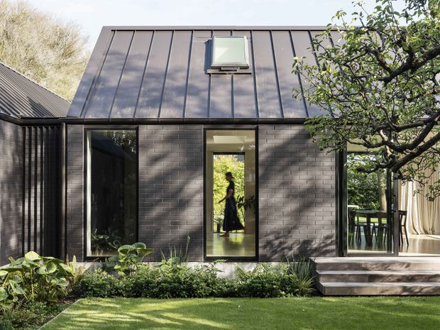 Exterior view of a compact contemporary extension or pavilion with dark brick walls and a standing-seam metal gable roof. Tall glazed openings connect the interior to a timber deck and landscaped garden, with a roof window and mature planting framing the facade.