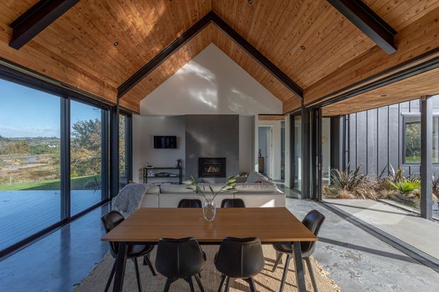 Open-plan contemporary living and dining area with a timber-lined vaulted ceiling, exposed dark structural beams, extensive black-framed glazing, and a strong connection to an outdoor terrace and landscape. A large timber dining table occupies the foreground, while a lounge and fireplace are positioned beyond.
