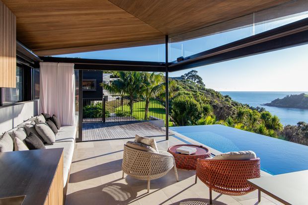 A luxurious open-plan indoor-outdoor living area opening onto a deck and infinity-edge pool, with panoramic coastal and ocean views. The space features a warm timber-lined ceiling, full-height glazing, dark slender frames, a pale sectional sofa, woven lounge chairs, and a round textured coffee table. Tropical landscaping and a rocky coastal headland are visible beyond the pool.