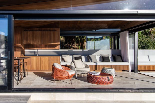 A contemporary indoor-outdoor living area viewed through large open black-framed glazing. The space features extensive warm timber wall and ceiling panelling, a built-in timber kitchenette or bar with stools, a long built-in window seat, and a light upholstered sofa. A pair of distinctive woven red-orange lounge chairs and a matching low round coffee table add colour. The room opens directly to a landscaped courtyard or garden and has a bright, relaxed yet refined atmosphere.