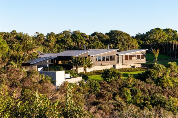 A low-profile single-storey contemporary house set within dense coastal vegetation. The elongated building has several connected wings, broad shallow-pitched metal roofs, extensive glazing, timber-toned wall cladding, and sheltered outdoor living areas. Landscaped terraces and native scrub surround the home, with a bright blue sky and mature trees in the background.