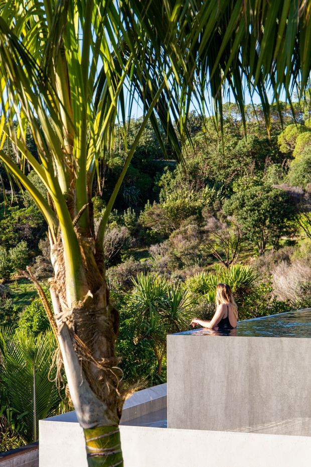 A sunlit outdoor pool area set within dense, landscaped vegetation. A raised, minimalist rectangular pool or spa with smooth pale concrete walls occupies the foreground, while a person sits partially submerged at its edge. Large tropical palm fronds dominate the view, with layered native-looking garden planting and a wooded hillside beyond.