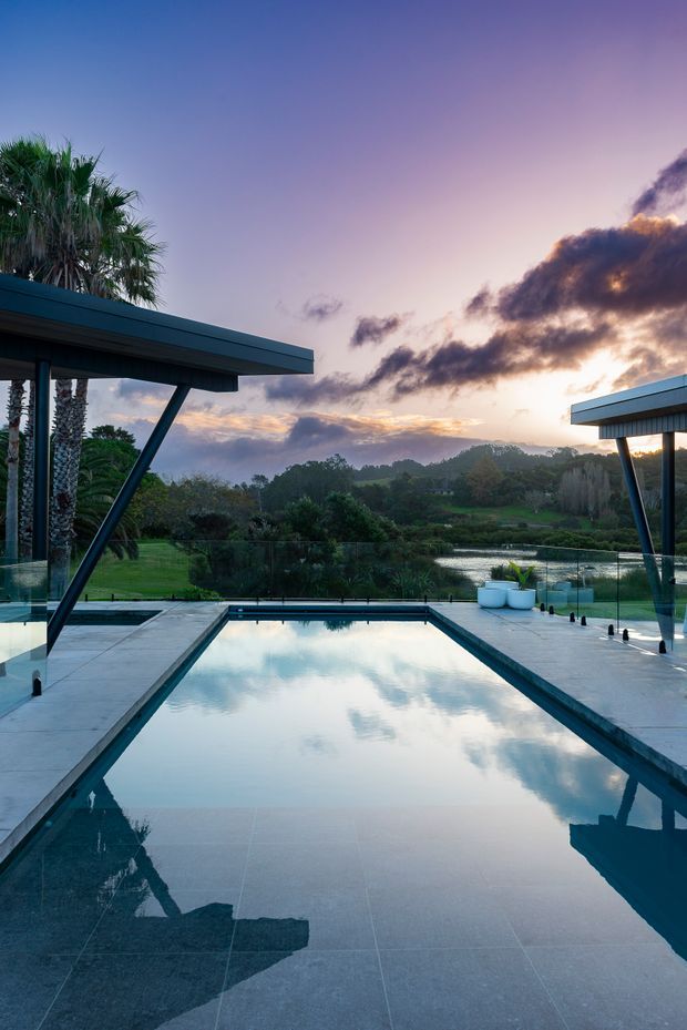 A tranquil modern outdoor pool terrace at sunset, with a long rectangular reflecting pool bordered by pale stone or concrete paving and frameless glass balustrades. Flat-roofed structures with dark metal framing and angled supports flank the pool, while lush tropical landscaping, palms, a pond, and rolling greenery extend into the background. The water reflects the dramatic pastel sky and surrounding architecture.