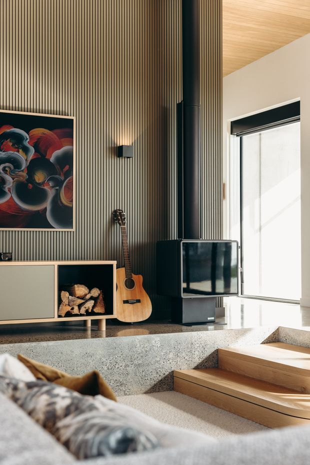 A warm, modern living room featuring a tall vertically slatted timber feature wall, a black wood-burning stove, television, guitar, and low timber media cabinetry with firewood storage. A sunlit window and timber-lined ceiling brighten the space, while polished concrete steps and a built-in seating area create a split-level arrangement. The foreground includes a large upholstered sofa.