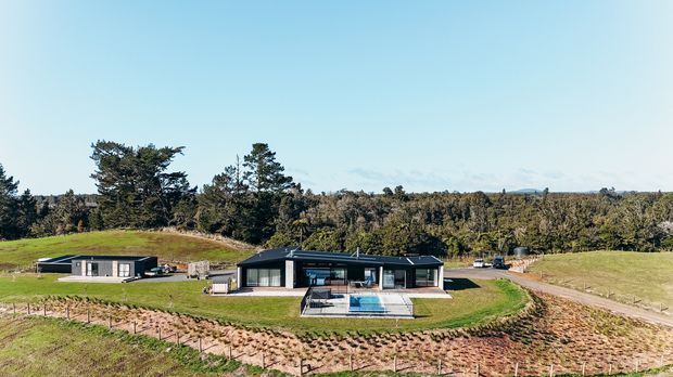 A low-profile contemporary single-storey home set in a spacious rural landscape, with dark cladding, expansive glazing, a broad roof form, and a fenced swimming pool in front. The property is surrounded by open grassed areas, cultivated earth, and dense native-looking woodland.