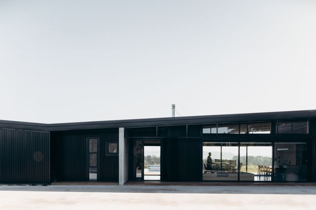 Single-storey contemporary building with a long, low flat roof and dark vertically corrugated exterior cladding. Large glazed openings reveal an interior living area and provide views toward a landscaped setting and water-like horizon. The facade has a recessed entry, narrow vertical windows, and broad floor-to-ceiling glazing, creating a minimalist linear composition.