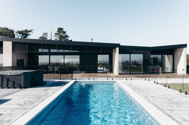 A modern single-storey residence viewed from the pool, featuring a long low-slung flat roof, dark metal cladding, expansive floor-to-ceiling glazing, and pale concrete paving. A rectangular swimming pool dominates the foreground, with landscaped surroundings and trees in the background.