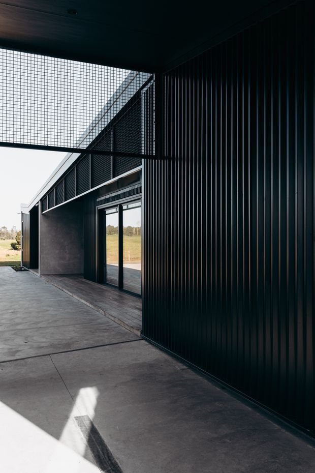 A dark, contemporary architectural exterior showing a covered concrete walkway beside a building clad in vertically ribbed black metal panels. Large glazed doors and windows open toward a grassy rural landscape, while a gridded overhead screen creates strong geometric shadows and filtered light.