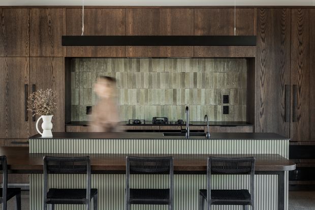 A refined, dark-toned kitchen with full-height vertical timber cabinetry, a large island with a fluted front and dark benchtop, three black bar stools, and a textured muted green-grey tiled splashback. A slim black rangehood and integrated cooking appliances sit within the cabinetry, creating a moody, contemporary interior.