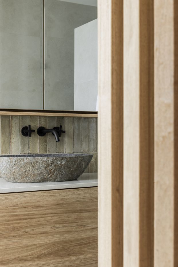 Close-up view of a contemporary bathroom vanity featuring a rough natural stone vessel basin, matte black wall-mounted tapware, a pale stone benchtop, warm timber cabinetry, and vertically ridged muted green-grey wall tiles. A large mirror and timber-framed opening complete the composition.
