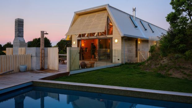 Exterior dusk view of a compact modern timber-clad house beside a swimming pool. The building has a steep gable roof with standing-seam metal roofing, roof skylights, extensive glazing, and an open illuminated living area. A timber deck, privacy fencing, landscaped lawn, and a sculptural outdoor chimney/fireplace structure occupy the foreground and side yard.