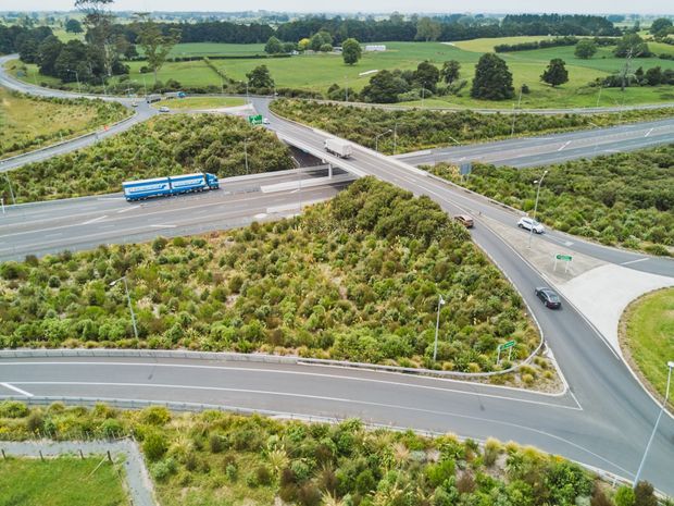 Aerial view of a large multilane highway interchange with overpasses, ramps, traffic signs, landscaped native vegetation, and surrounding green rural farmland.