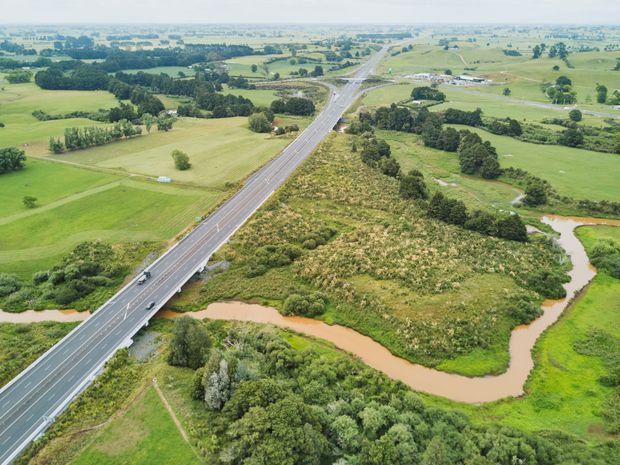 Aerial view of a rural landscape featuring a divided roadway and bridge crossing a winding muddy-brown river or creek, surrounded by green farmland, scattered trees, hedgerows, and distant agricultural fields.