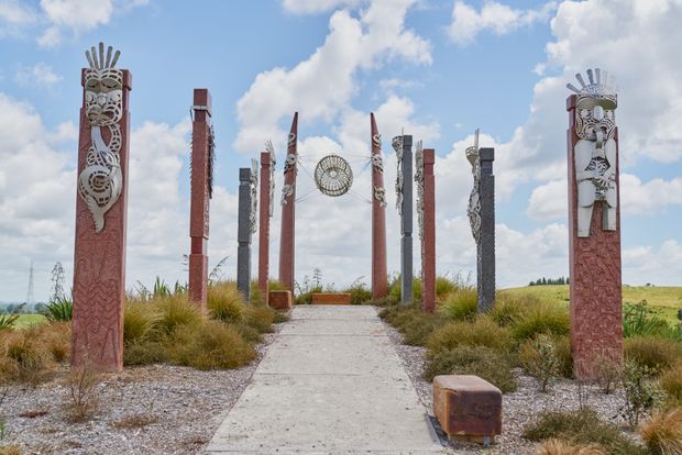 A landscaped outdoor ceremonial walkway lined with tall carved red and dark sculptural columns, including intricate Māori-inspired motifs and suspended circular artwork. The concrete path passes through native grasses under a bright blue sky, with open rolling countryside in the background.