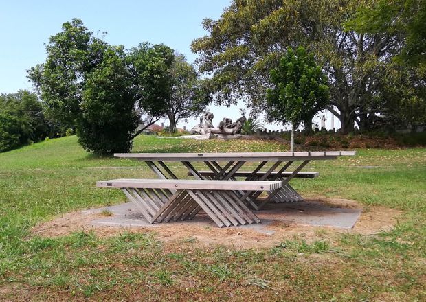 Outdoor photograph of a weathered timber picnic table with attached bench seating, positioned on a small concrete pad in a grassy, tree-lined park or garden setting. A sculptural feature and mature landscaping are visible in the background under clear daylight.