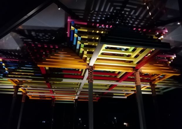 A dramatic nighttime view looking upward at a large illuminated overhead architectural installation or canopy. Layered horizontal slats and beams form a dense geometric ceiling in vivid yellow, orange, red, blue, green, and white, supported by slender dark columns with patterned detailing. Strong shadows and reflections create a highly graphic, immersive composition.