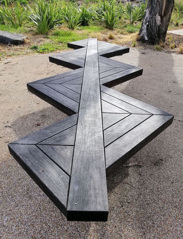 Outdoor landscaped setting featuring a striking geometric dark timber walkway or sculptural deck element laid across a gravel or compacted aggregate surface. The structure consists of angular zigzagging platforms with visible plank joints and a raised central spine, surrounded by spiky ornamental planting and mature trees.